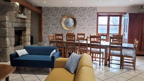 A living room with a stone fireplace, a dining area with wooden chairs and a table, and a large window. - Gîte du Platane, Sleeps 14 (Grazac)