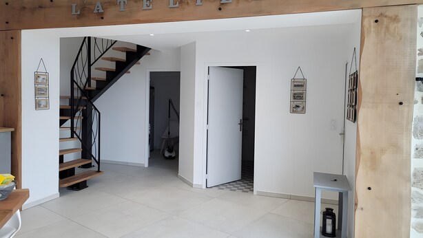 Un interior moderno con una escalera, una viga de madera con el letrero 'L'ATELIER' y una pequeña mesa con un farol.