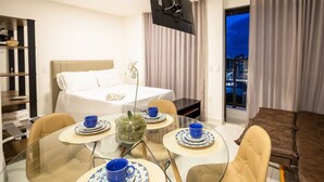 A hotel room with a bed, a dining table with blue cups and plates, a glass coffee table with a plant, and a city view through the window. - Compact Tambau #704 - Apartment By Carpediem (João Pessoa)