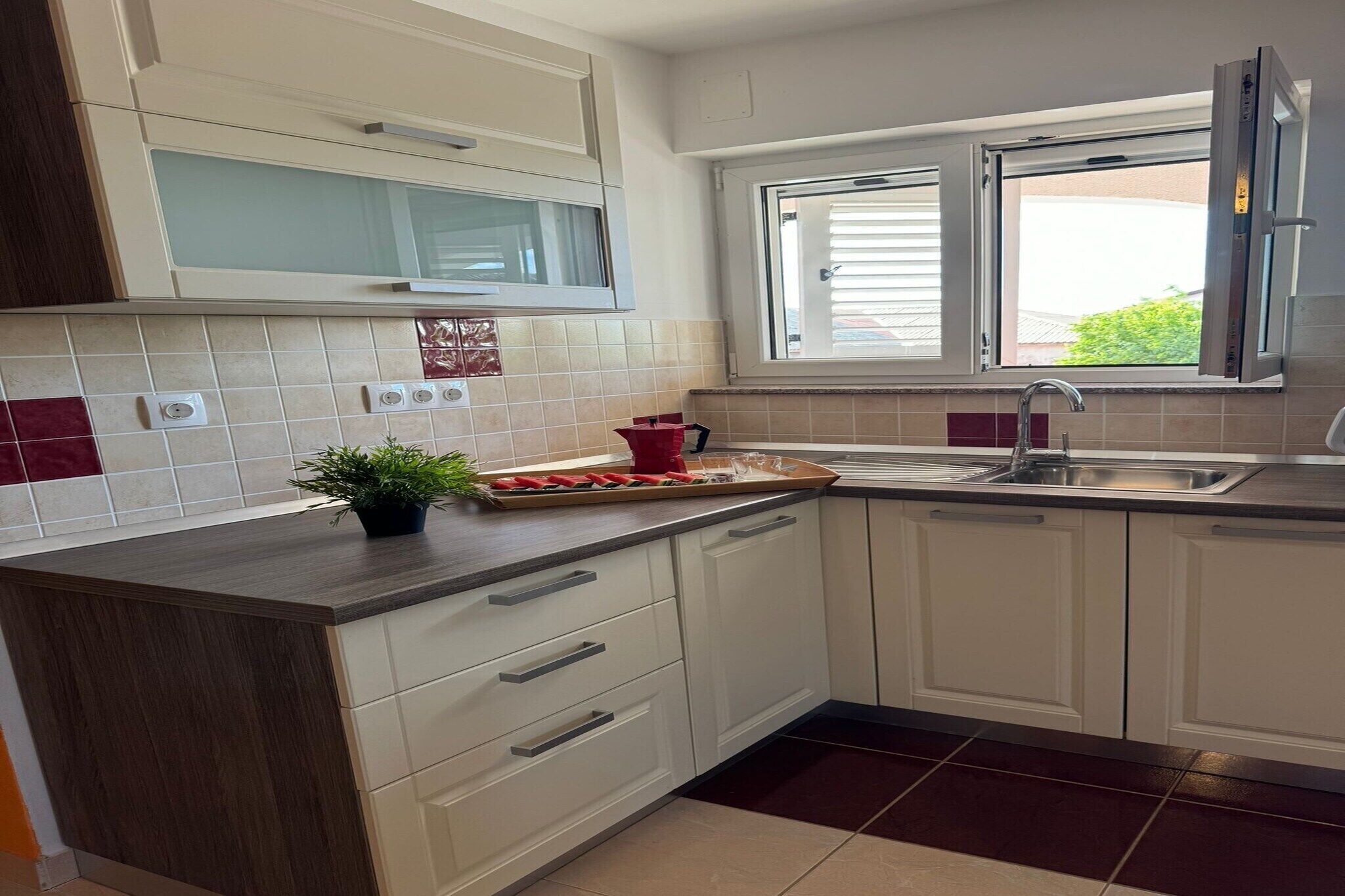 Une cuisine moderne avec des meubles blancs, un plan de travail foncé, une fenêtre avec des stores et une plante en pot posée sur le plan de travail.