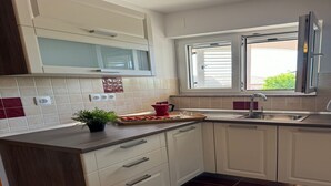 Une cuisine moderne avec des meubles blancs, un plan de travail foncé, une fenêtre avec des stores et une plante en pot posée sur le plan de travail.