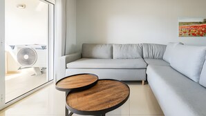 Un salon moderne avec un canapé d’angle gris, deux petites tables rondes en bois, une grande fenêtre donnant sur un champ et un ventilateur de plafond.