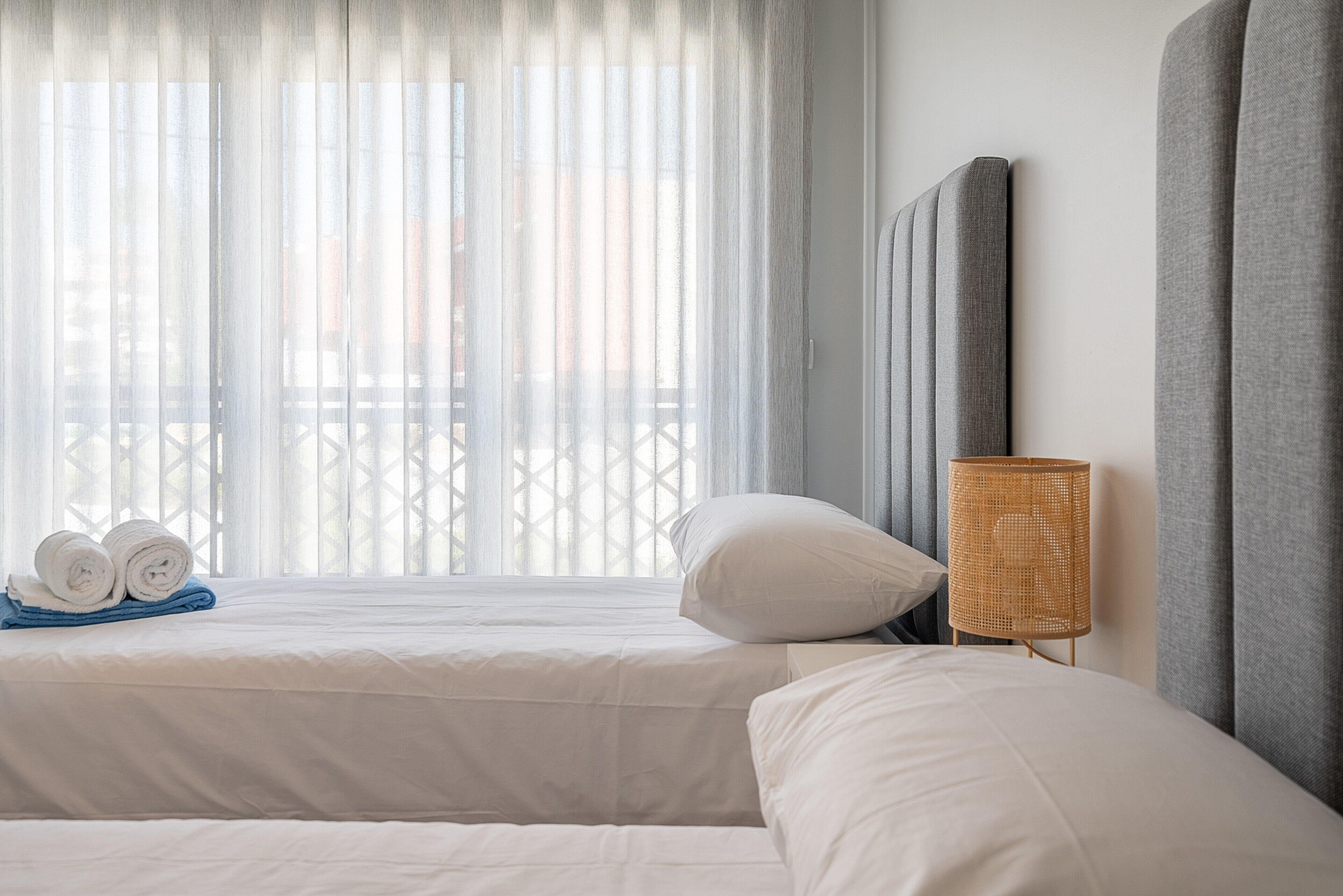 Een hotelkamer met twee bedden, een groot raam met dunne gordijnen, een rieten lamp en uitzicht op de omgeving.