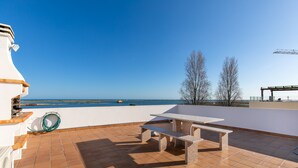 Um terraço com mesa e bancos, com vista para a água e árvores.