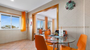 Uma sala de jantar com mesa de vidro, cadeiras laranja e vista através de uma porta aberta.