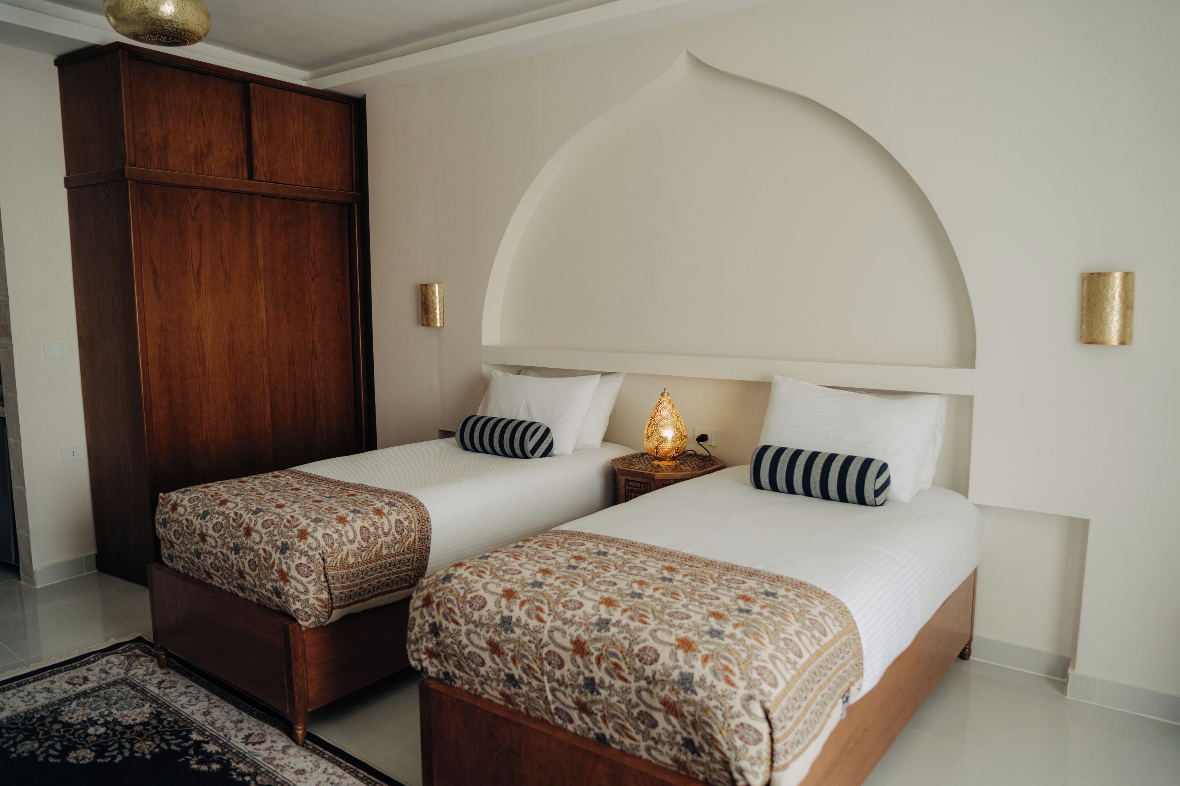 Hotelkamer met twee bedden, een garderobe, een nachtkastje met lamp en een decoratieve boog in de muur.