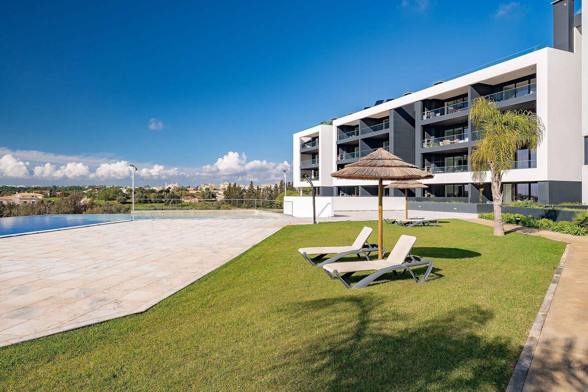Un edificio moderno con piscina, sillones de descanso y una sombrilla de paja.