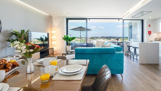A modern living room with a large window, a teal sofa, a dining area with breakfast setup, and a kitchen area. - Luxury 2-Bedroom Apartment with Sea View and Pool (Portimão)