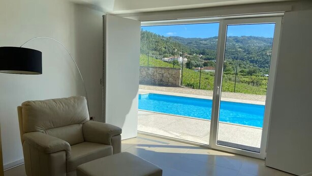 A room with a beige sofa, a matching ottoman, and a large sliding glass door leading to a pool. - Enjoy our heated swimming pool, both in summer and winter. (Ponte de Lima)