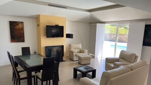 A modern living room with a dining area, a flat-screen TV mounted on the wall, a fireplace, and a large window offering a view of the outdoors. - Enjoy our heated swimming pool, both in summer and winter. (Ponte de Lima)
