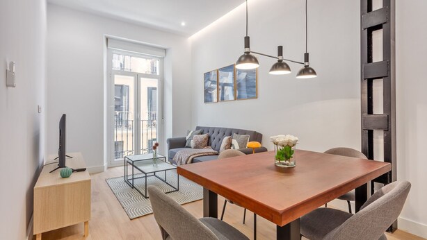 A modern living room with a wooden dining table, grey sofas, and a large window with a view of buildings. - Cozy 1BR Retreat Steps from Sol & Santa Ana (Madrid)