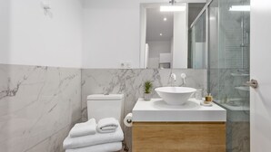 A modern bathroom with a marble backsplash, a white sink, a toilet, and a glass shower enclosure. - Cozy 1BR Retreat Steps from Sol & Santa Ana (Madrid)