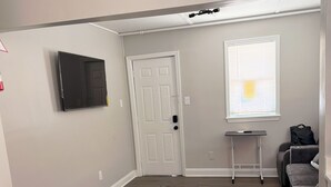 Un ambiente compatto con una TV a schermo piatto montata a parete, una porta con maniglia, un tavolino, una sedia e un divano grigio.