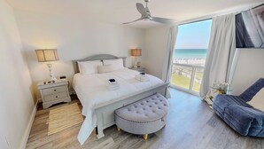 Un dormitorio con cama, mesitas de noche, ventilador de techo, vista a la playa y una silla.