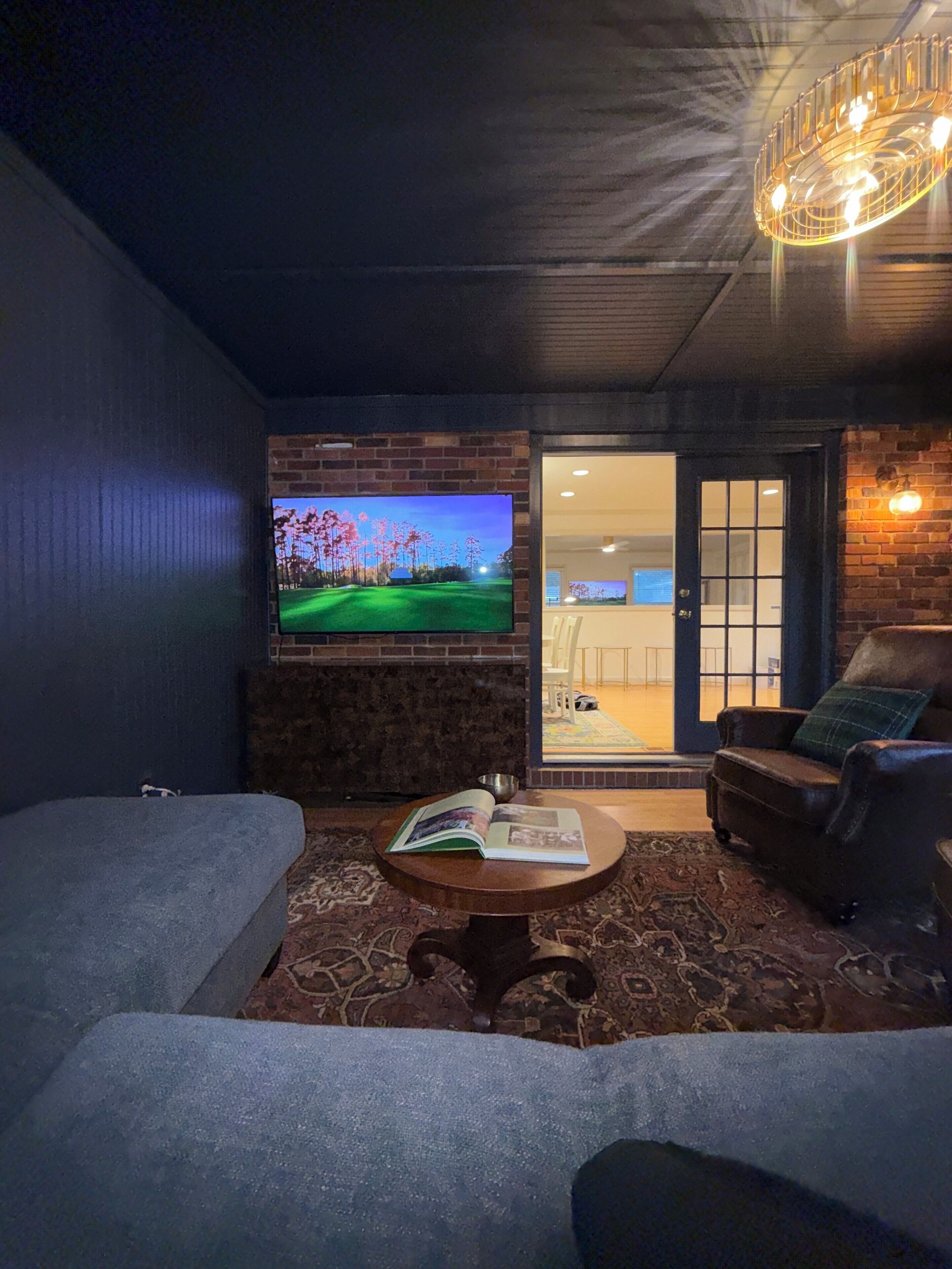 A cozy living room with a flat-screen TV mounted on a brick wall, a comfortable sofa, a coffee table with a book, and a view of the outside through a glass door.
