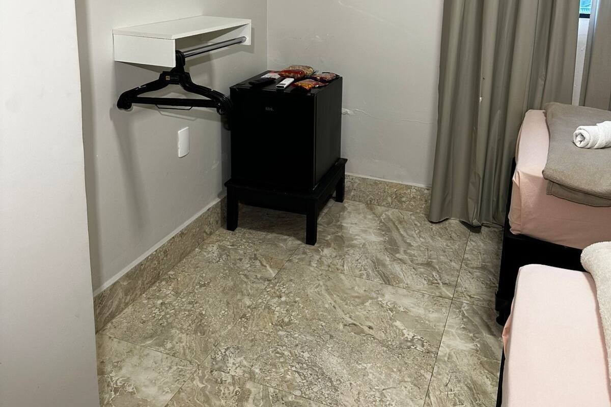Habitación de hotel con televisor de pantalla plana montado en la pared, un mueble negro pequeño, una toalla sobre la cama y una ventana con cortinas.
