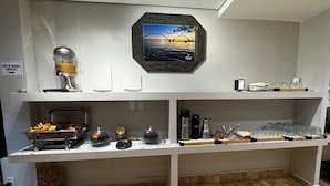 Una barra de desayuno con una cafetera, un cartel que dice 'Café da Manhã Cortesia' y una foto enmarcada de un atardecer sobre el agua.