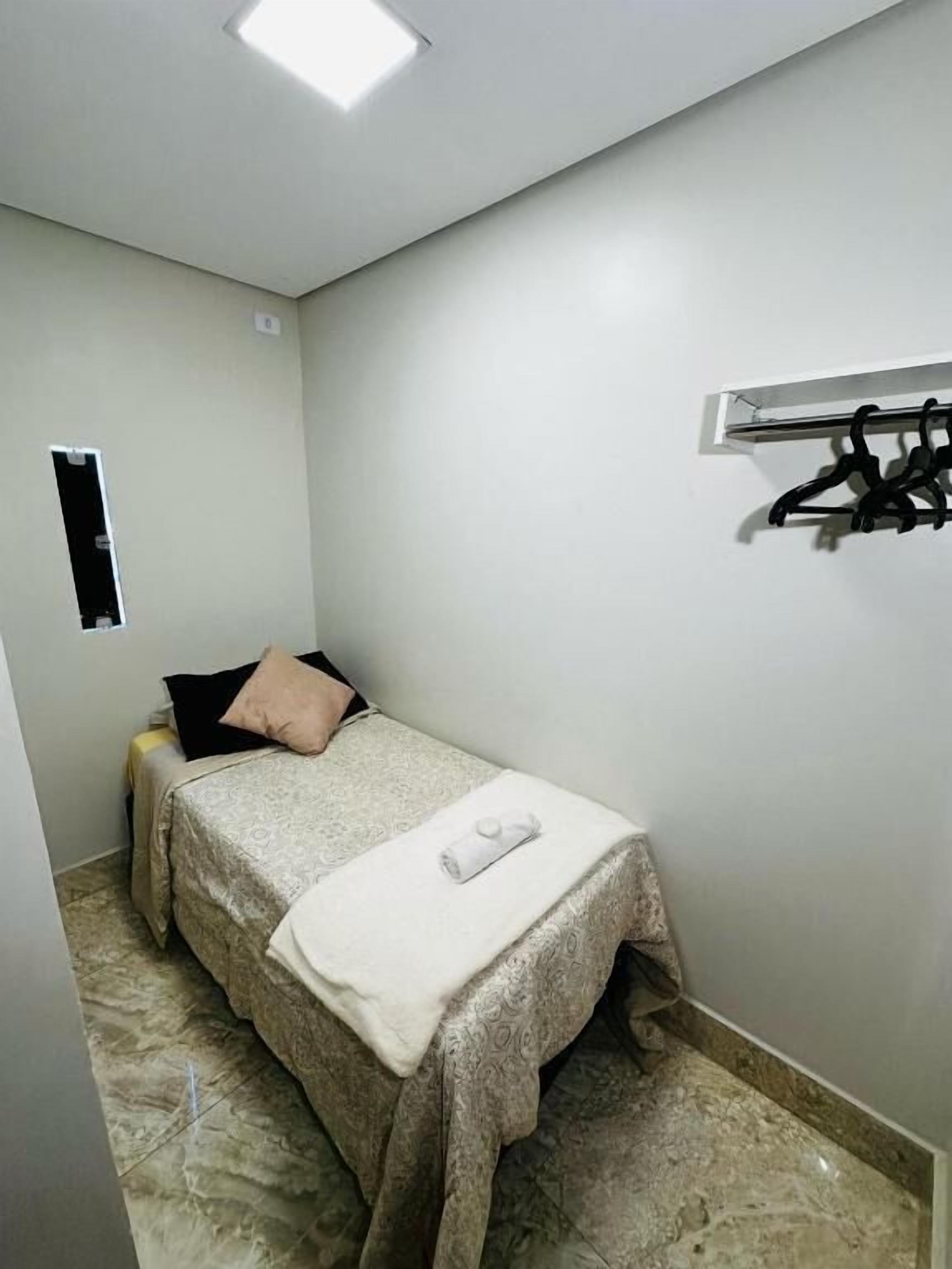 Une petite chambre à coucher, bien rangée, avec un lit, une table de chevet, un porte-manteau et une petite fenêtre.