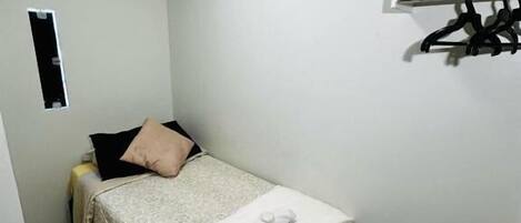 Une petite chambre à coucher, bien rangée, avec un lit, une table de chevet, un porte-manteau et une petite fenêtre.