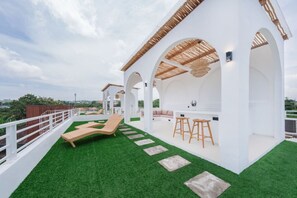 A rooftop terrace with artificial grass, a lounge chair, a bar area with stools, and white arches.