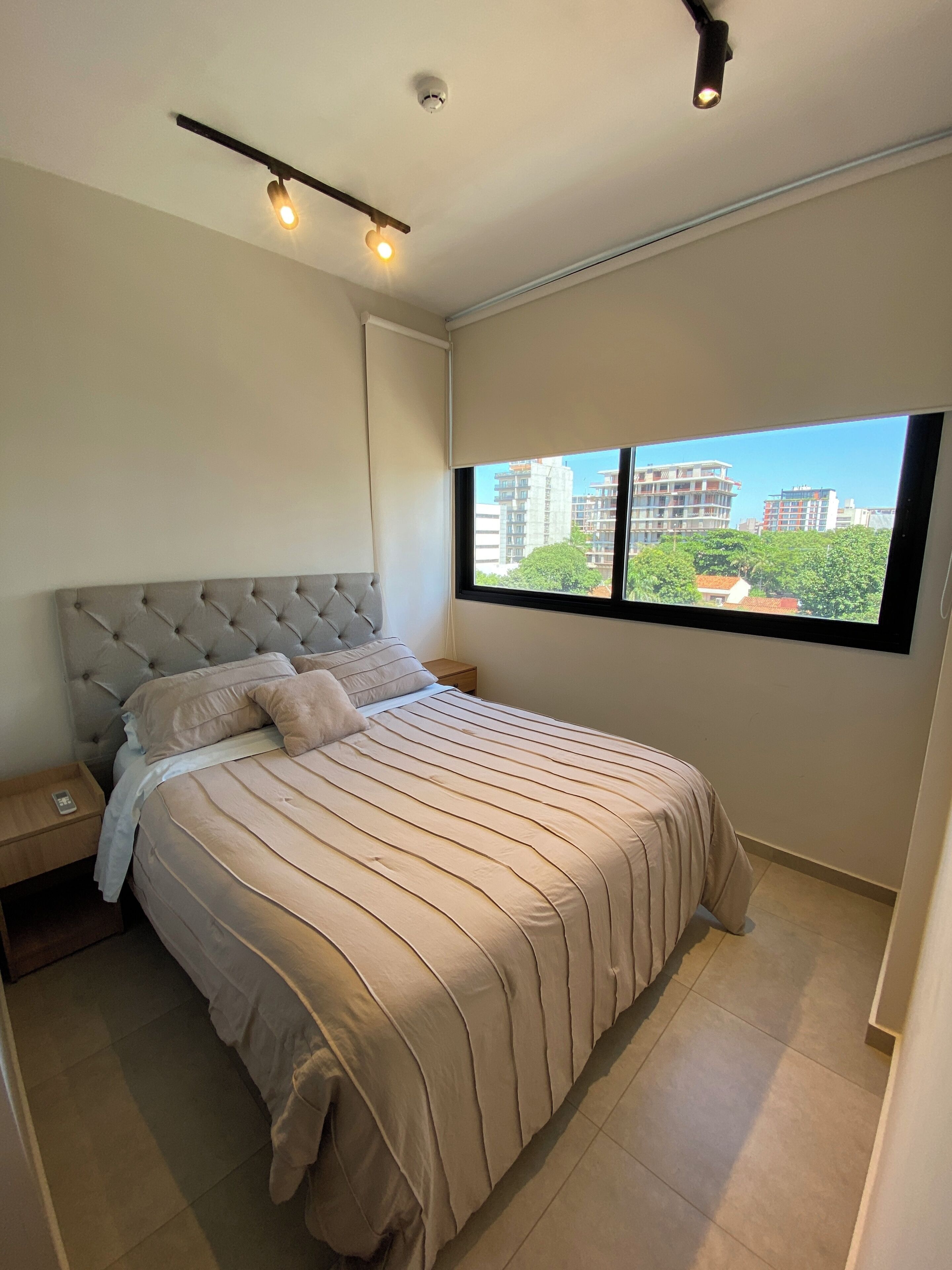 Un dormitorio con una cama grande, una mesita de noche, un ventanal con vistas a edificios y una lámpara en el techo.