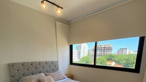 Un dormitorio con una cama grande, una mesita de noche, un ventanal con vistas a edificios y una lámpara en el techo.