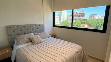 Un dormitorio con una cama grande, una mesita de noche, un ventanal con vistas a edificios y una lámpara en el techo.