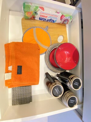 A drawer containing a stack of orange towels, a patterned cloth, a wooden tray with a checkered cloth, a red bowl, a red plate, and several black and white striped bottles.