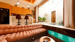 A modern living room with a large cushioned sofa, a small round table, and a view of a pool through a glass wall. - Modern 1BR Oasis - Sunken Lounge & Near Canggu (Mengwi)