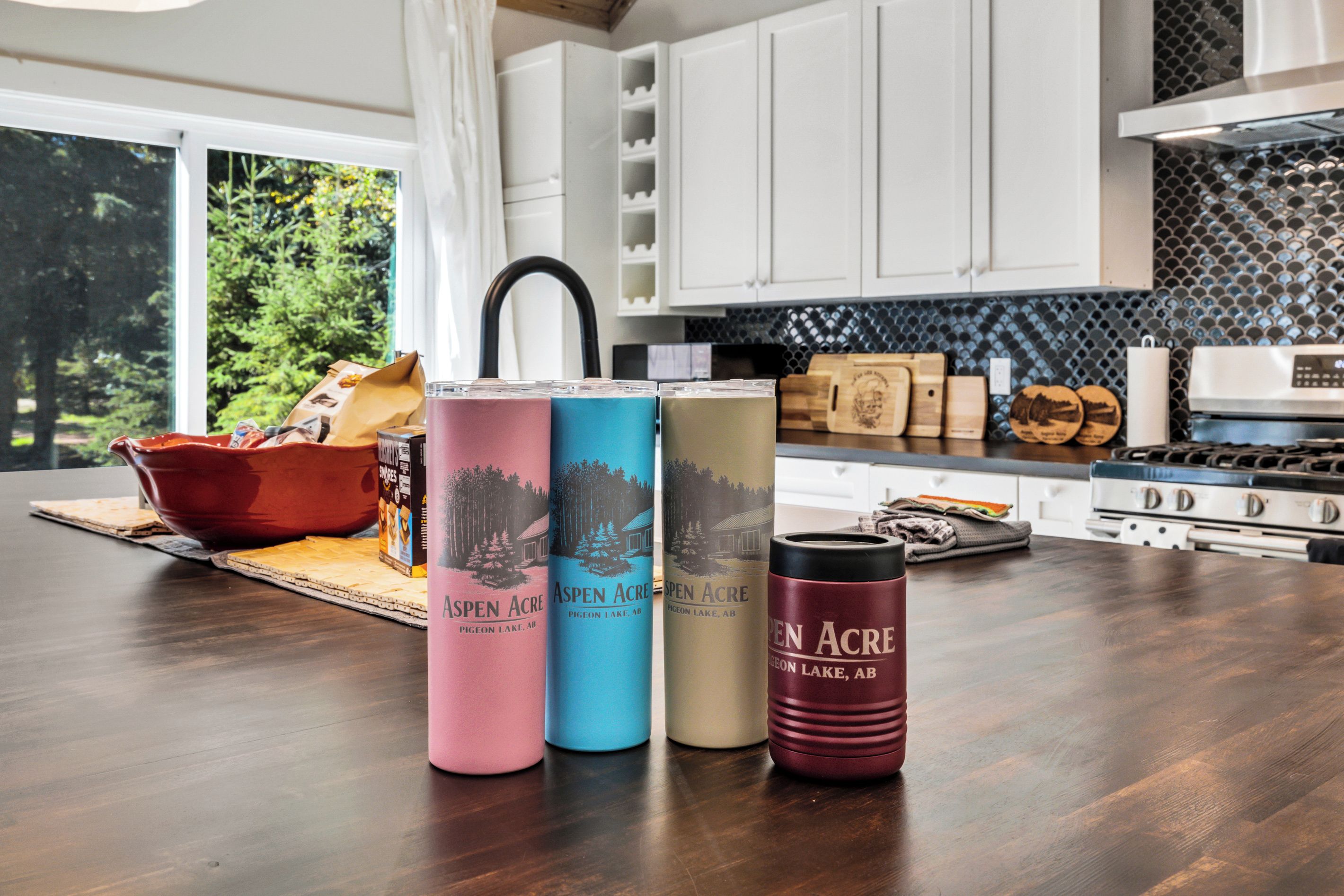 A kitchen with white cabinets, a black backsplash, and a wooden countertop. There are three beverage containers labeled 'Aspen Acre' and a red mug.