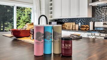 A kitchen with white cabinets, a black backsplash, and a wooden countertop. There are three beverage containers labeled 'Aspen Acre' and a red mug.