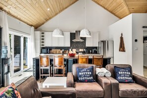 Une cuisine moderne avec des armoires blanches, un pare-feu noir et un réfrigérateur en acier inoxydable. Deux luminaires suspendus sont placés au-dessus de l’îlot central, et le plafond est en bois.