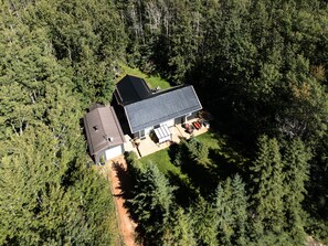Une maison à deux étages dotée d’une grande terrasse et d’un garage, entourée d’une forêt dense.