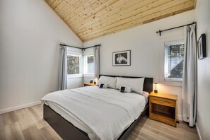 Une chambre à coucher avec un lit, des tables de chevet, une commode, une fenêtre avec des rideaux, un tableau au mur et un plafond en bois.