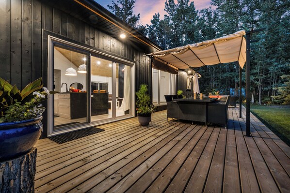 A wooden deck with a covered outdoor seating area and a sliding glass door leading to an interior space with a kitchen and dining area.