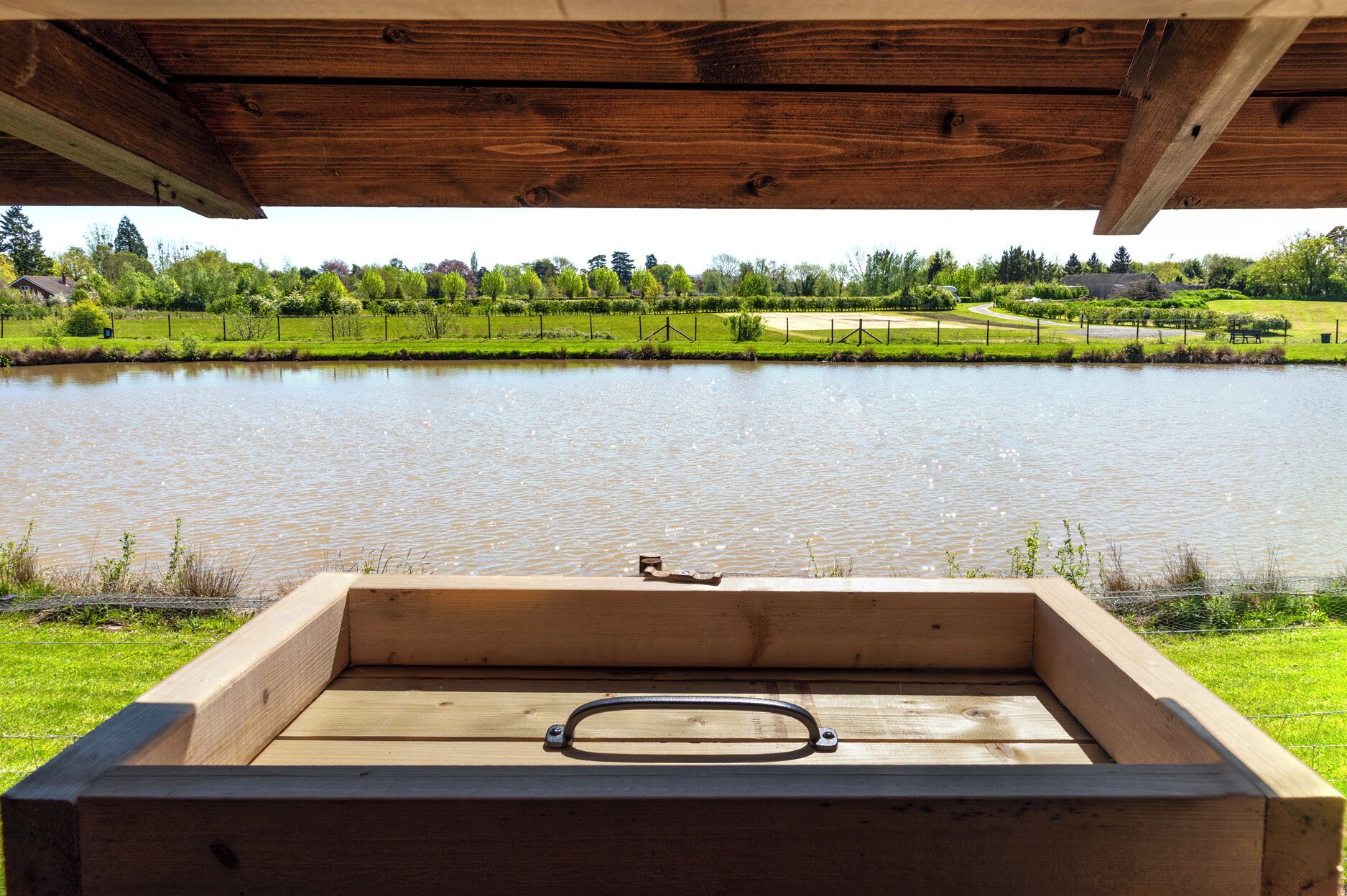 Un banco de madera con un asa de metal, con vista a un lago tranquilo y un paisaje cubierto de hierba, con árboles al fondo.
