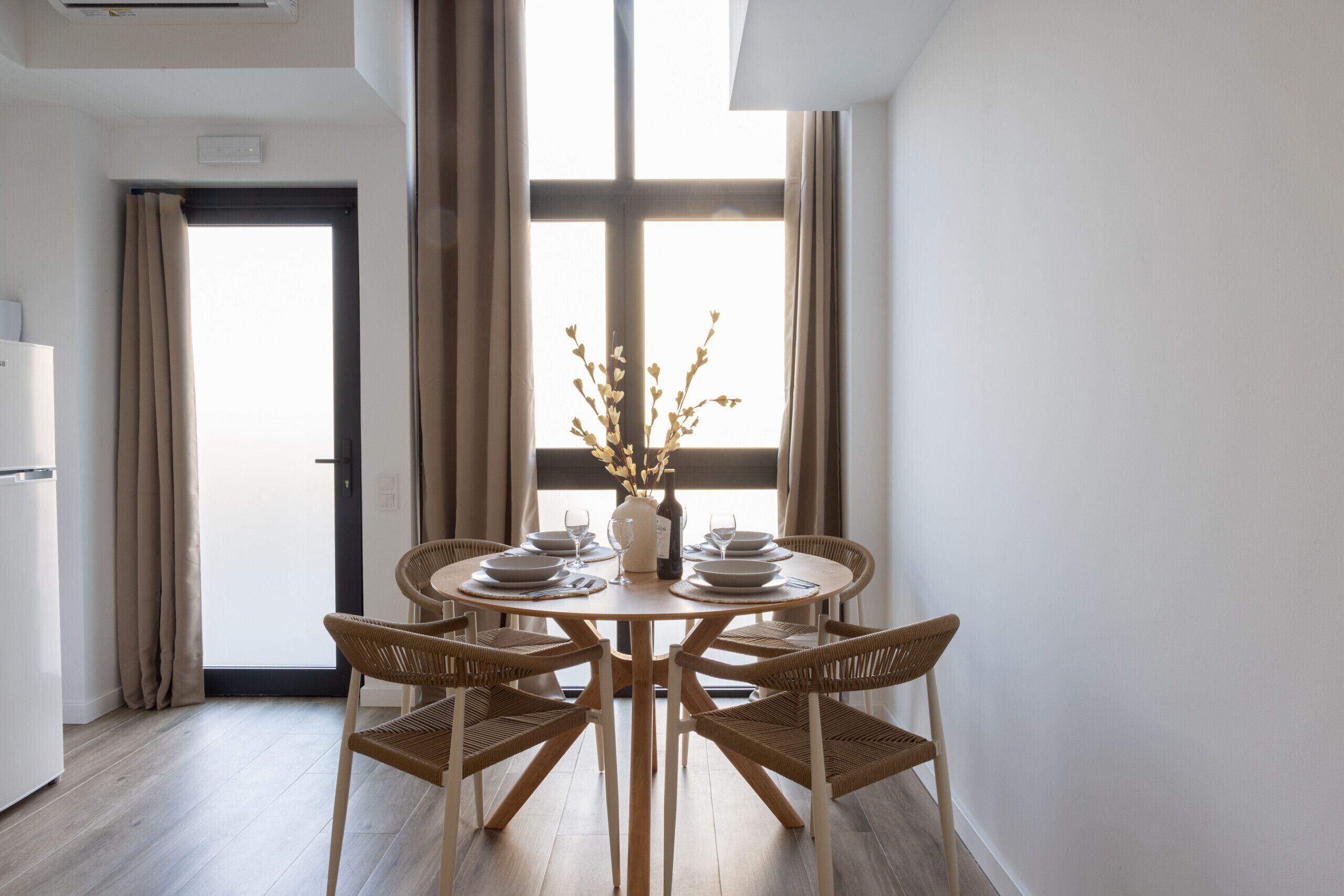 Een eetruimte met een ronde tafel, vier rieten stoelen en een vaas met takken.