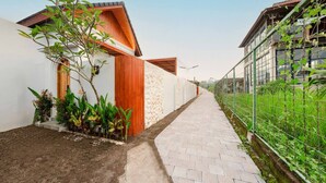 A narrow pathway flanked by buildings with a mix of modern and traditional architecture, a small garden area with plants, and a metal fence on one side. - Stylish 2BR Villa near Central Ubud (Ubud)