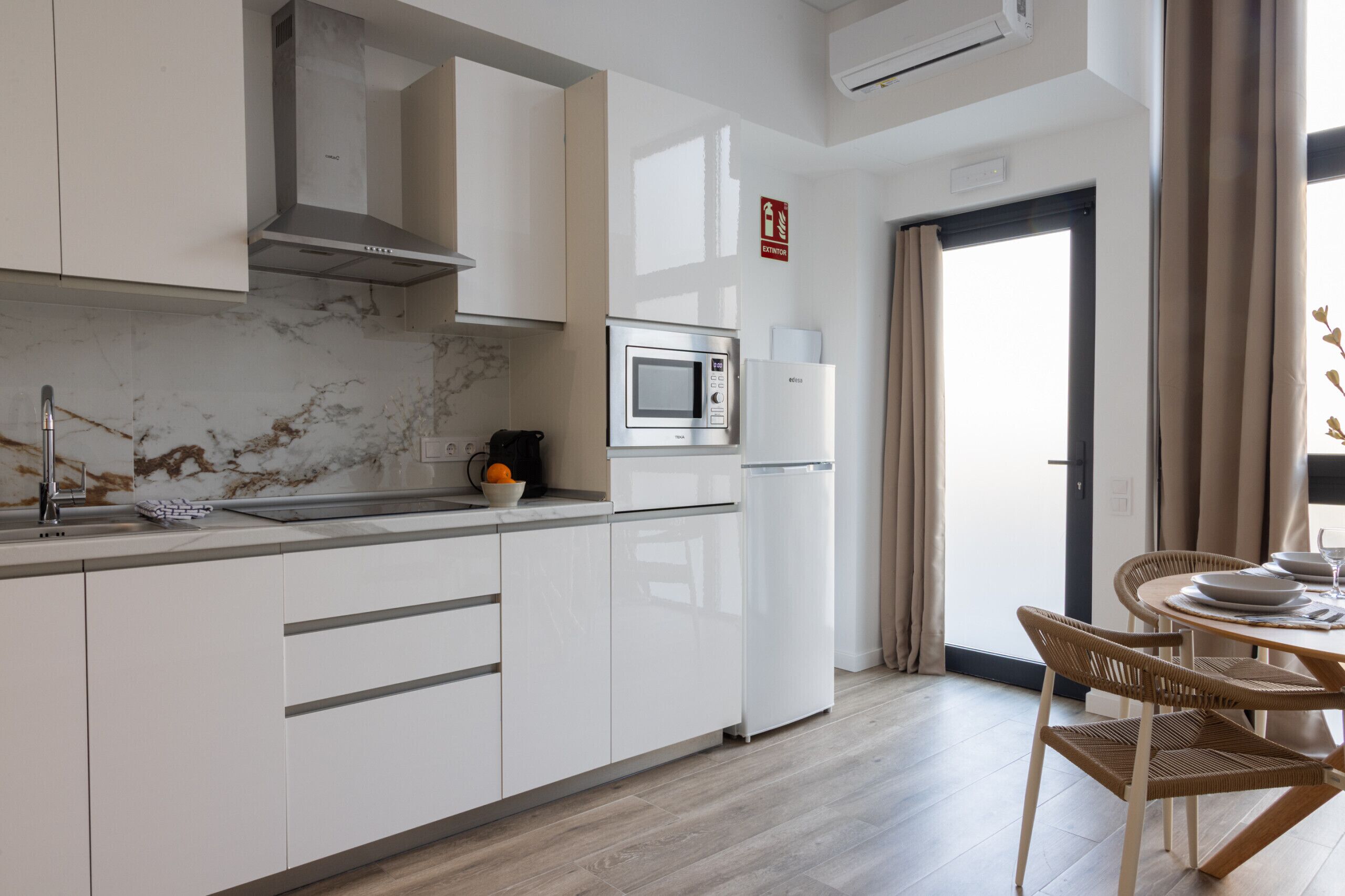Een moderne keuken met witte kasten, een ingebouwde magnetron en een koelkast. Er is een eethoek met een tafel en stoelen.