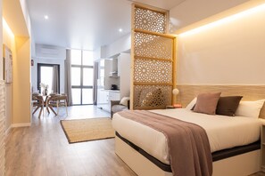 Ein modernes Schlafzimmer mit einem großen Bett, einem dekorativen Holzschirm, einer Essecke mit Stühlen und Tisch sowie einer Küchenzeile im Hintergrund.
