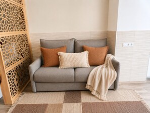 Ein graues Sofa mit zwei braunen und einer beigen Kissen, einer beigen Tagesdecke und einem gemusterten Teppich.