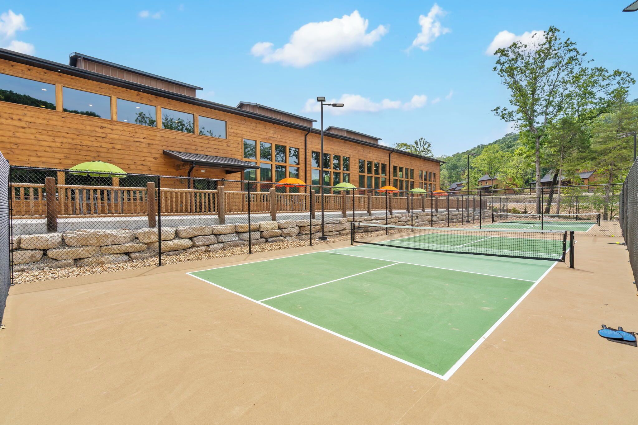 An outdoor tennis court with a wooden fence, a building with large windows, and outdoor seating with umbrellas.