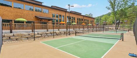 An outdoor tennis court with a wooden fence, a building with large windows, and outdoor seating with umbrellas.