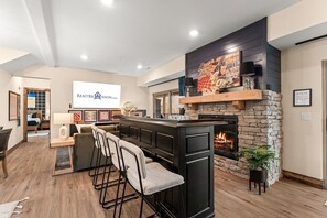 Uma sala de estar moderna com lareira de pedra, área de bar com banquetas e uma tela grande exibindo um logotipo.