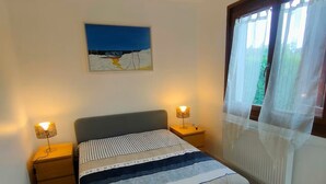 A bedroom with a bed, two bedside tables, a lamp, a painting, and a window with curtains. - Charming house in a quiet location, in the center of town, less than 800 m from the beach and all amenities (Cabourg)