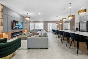 A modern living room with a flat-screen TV, a fireplace, a sofa, and a dining area with bar stools. - 10BR house with pool table near Disney area (Orlando)