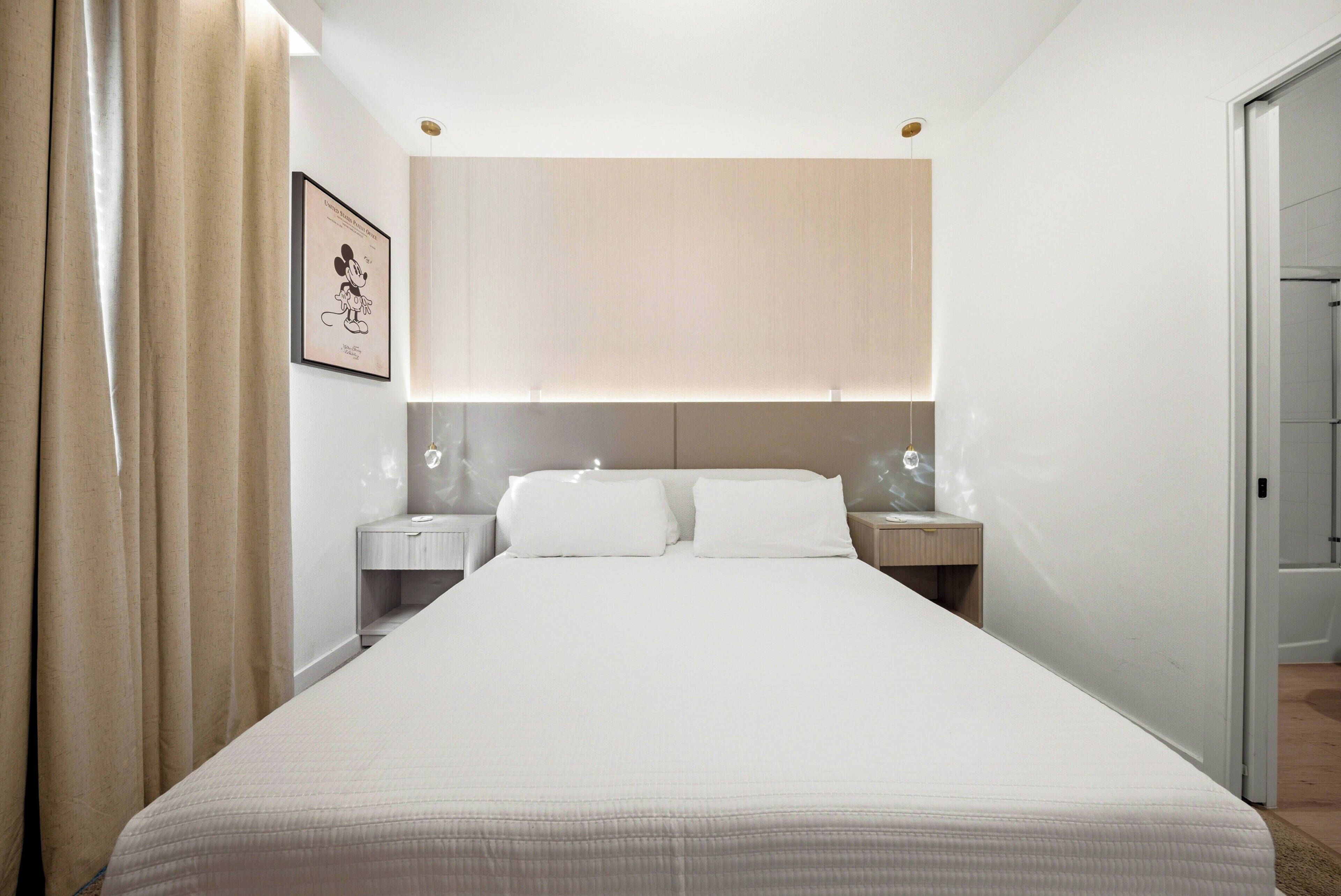 Welcome to your cozy retreat, featuring a comfortable double bed with crisp white linens. Soft beige accents and warm lighting create a soothing vibe, enhanced by elegant drapes and tasteful wall art for an inviting atmosphere.