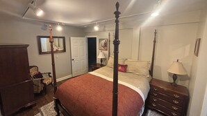 Een hemelbed met een rode spreideken, een houten nachtkastje met een lamp, een kast en een stoel met een gekleurd kussen.