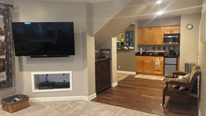 Een woonkamer met een flatscreen televisie, een houten kast en een keuken met houten keukenkastjes en een magnetron.