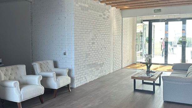 A spacious lobby with a high ceiling, exposed wooden beams, white brick walls, and a seating area with white upholstered chairs. - Tudcon Palm (Madrid)